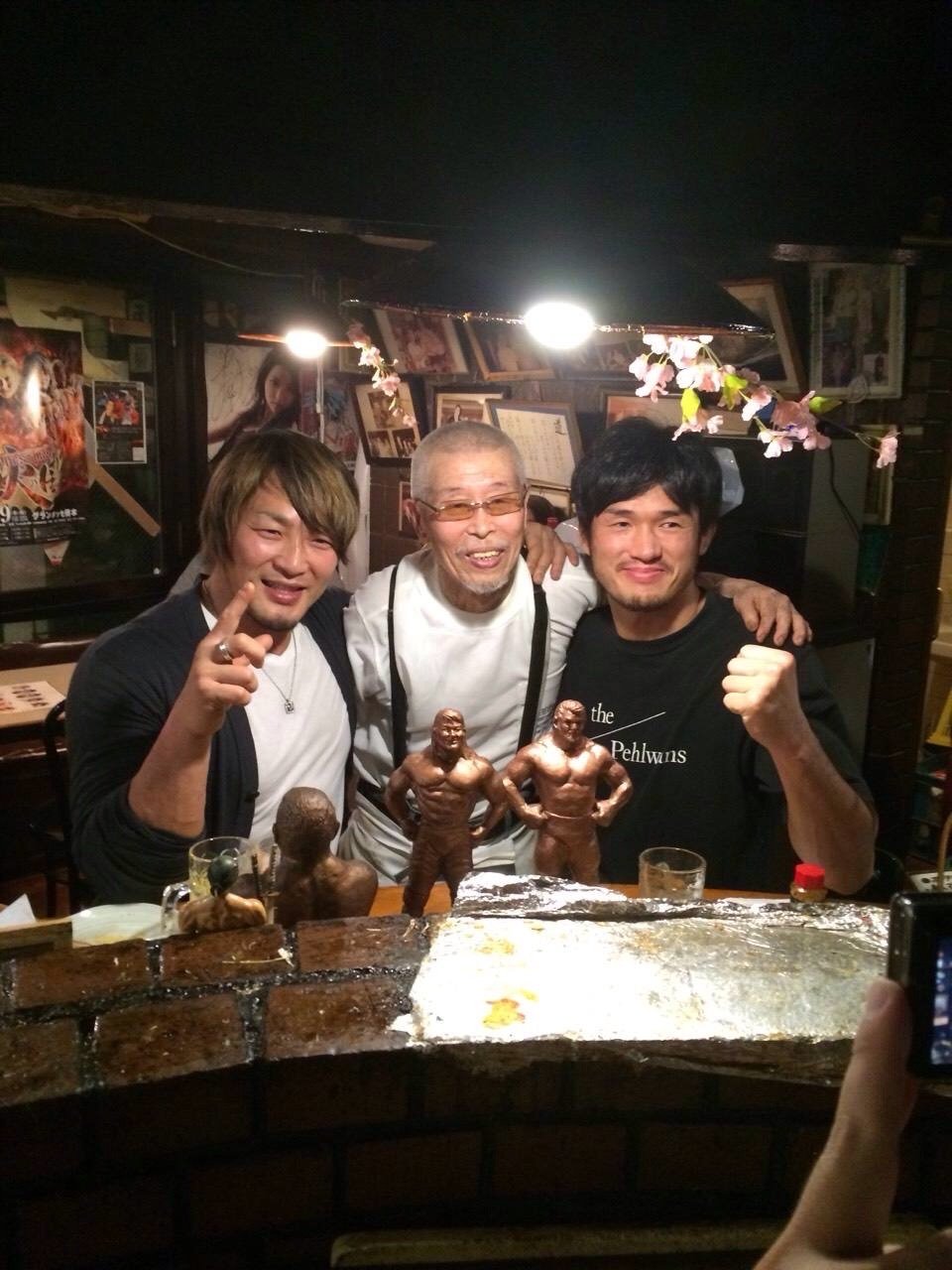 ブロンズ像を前に村田マスターと二人のレスラーが肩を組む様子